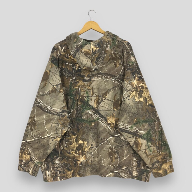 Op de afbeelding: Een camouflage hoodie met een bospatroon in bruin, groen en beige. De hoodie hangt aan een houten hanger tegen een lichtgrijze achtergrond. Het heeft een capuchon met trekkoord en een kangoeroezak.