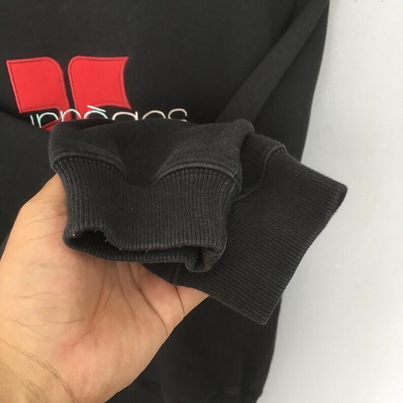 May include: Black sweatshirt with red and white embroidered logo. The logo features two red shapes and white text. The ribbed cuff of the sleeve is visible. The sweatshirt is made of a soft, comfortable material.