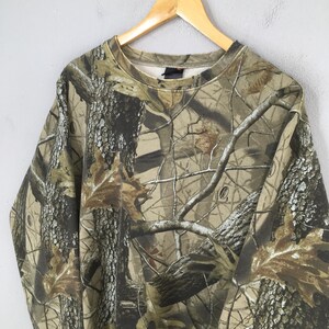 Puede incluir: Sudadera de manga larga con estampado de camuflaje. El dise&ntilde;o presenta una mezcla de colores marr&oacute;n, verde y beige, imitando un entorno forestal. La sudadera est&aacute; colgada en una percha de madera sobre un fondo gris claro.