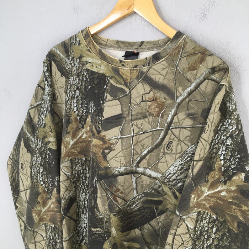 Puede incluir: Sudadera de manga larga con estampado de camuflaje. El dise&ntilde;o presenta una mezcla de colores marr&oacute;n, verde y beige, imitando un entorno forestal. La sudadera est&aacute; colgada en una percha de madera sobre un fondo gris claro.