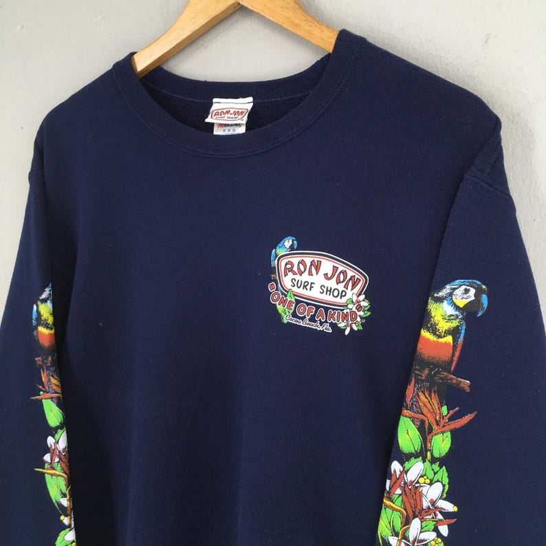 Puede incluir: Sudadera azul marino con el logotipo de Ron Jon Surf Shop y gr&aacute;ficos de loros en las mangas. El logotipo presenta un loro y las palabras "Ron Jon Surf Shop" y "One of a Kind".