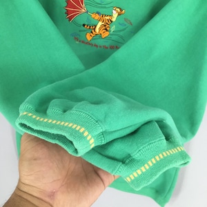 May include: Green sweatshirt featuring a graphic of Tigger from Winnie the Pooh holding an umbrella. The sweatshirt has yellow and white striped cuffs and the text "It's a blustery day in The 100 Acre Wood."