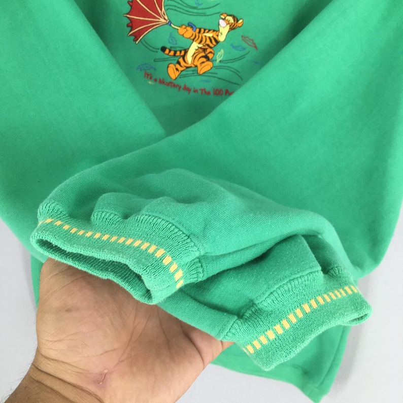 May include: Green sweatshirt featuring a graphic of Tigger from Winnie the Pooh holding an umbrella. The sweatshirt has yellow and white striped cuffs and the text "It's a blustery day in The 100 Acre Wood."