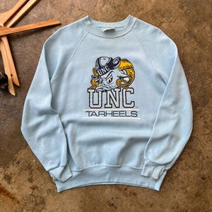 May include: A light blue crewneck sweatshirt featuring a graphic of a ram wearing a hat with the text UNC above it. Below the ram, the text UNC is printed in large block letters, followed by the word TARHEELS underlined by two horizontal lines.