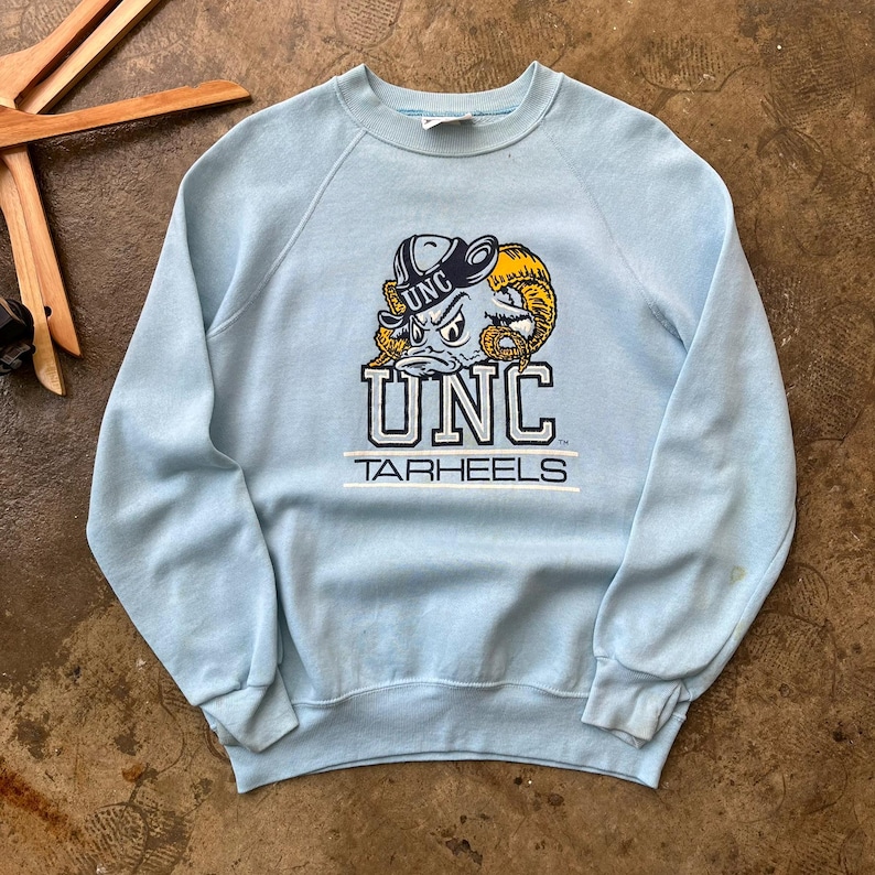 May include: A light blue crewneck sweatshirt featuring a graphic of a ram wearing a hat with the text UNC above it. Below the ram, the text UNC is printed in large block letters, followed by the word TARHEELS underlined by two horizontal lines.
