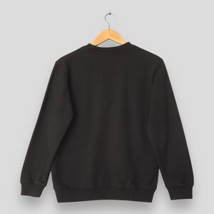 May include: A black crew neck sweatshirt hanging on a wooden hanger. The sweatshirt has long sleeves and a ribbed hem and cuffs. The background is a solid light gray.