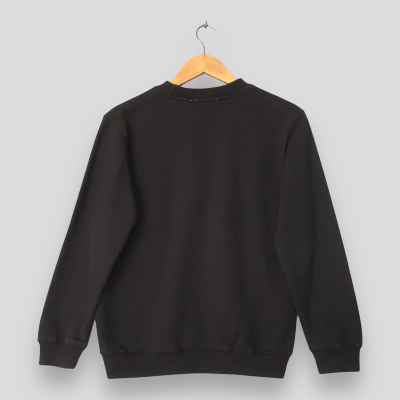 May include: A black crew neck sweatshirt hanging on a wooden hanger. The sweatshirt has long sleeves and a ribbed hem and cuffs. The background is a solid light gray.