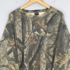 Op de afbeelding: Een sweatshirt met lange mouwen en een ronde hals, voorzien van een realistisch bos-camouflagepatroon in tinten bruin, beige en donkergroen. De stof toont een gedetailleerde print van boomstammen, takken en gevallen eikenbladeren over het hele kledingstuk.
