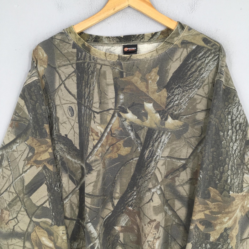 Op de afbeelding: Een sweatshirt met lange mouwen en een ronde hals, voorzien van een realistisch bos-camouflagepatroon in tinten bruin, beige en donkergroen. De stof toont een gedetailleerde print van boomstammen, takken en gevallen eikenbladeren over het hele kledingstuk.