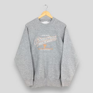 Puede incluir: Una sudadera gris con el texto "1998 SEC Champions Back to Back Volunteers" en letras naranjas y blancas. La sudadera está colgada de una percha de madera.