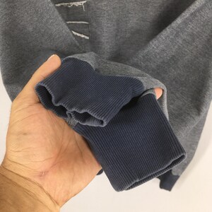 May include: Close-up of a gray sweatshirt sleeve with a navy blue ribbed cuff. The sleeve has a denim-like patch at the elbow. The fabric appears to be a soft, comfortable material.