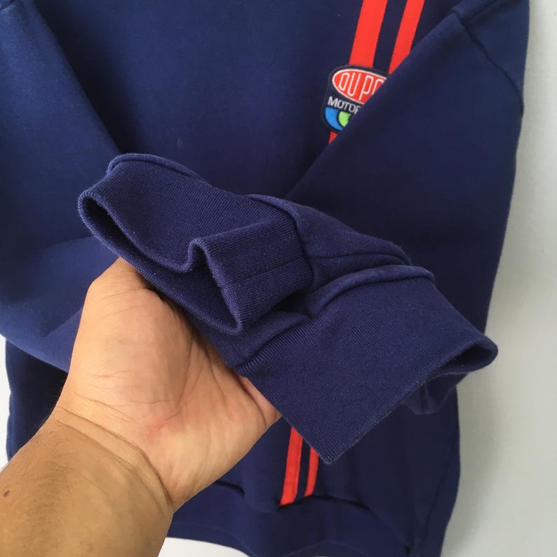 May include: A navy blue sweatshirt with red stripes and a logo. The sweatshirt has a ribbed cuff and a small logo patch. The red stripes are vertical and run down the sleeve. The logo is a circular patch with text.