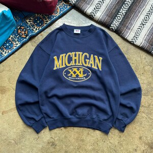 May include: A navy blue crewneck sweatshirt featuring the word MICHIGAN in yellow arched lettering above an oval graphic containing the text XXL and WOLVERINES in yellow. The garment has long sleeves, a ribbed collar, and ribbed cuffs.