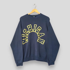 May include: A navy blue crewneck sweatshirt featuring the word MICHIGAN printed in an arched, collegiate-style font. The lettering is yellow with a dark blue outline. The garment has long sleeves, a ribbed collar, and ribbed cuffs.