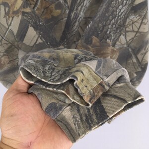 Op de afbeelding: Een sweatshirt met lange mouwen voorzien van een realistisch bos-camouflagepatroon in tinten bruin, beige, grijs en donkergroen. Het kledingstuk heeft geribbelde gebreide boorden bij de polsen en een bijpassende camouflageprint over de gehele stof.