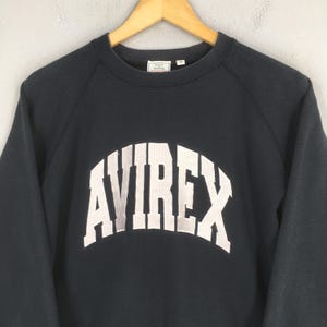 May include: A dark navy blue long-sleeve sweatshirt featuring the word AVIREX in large, arched, light gray block lettering across the chest. The garment has a crew neckline and a small white interior brand label at the back of the neck.