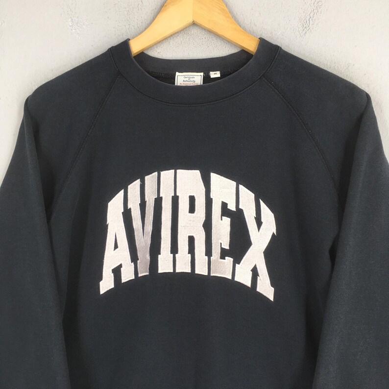 May include: A dark navy blue long-sleeve sweatshirt featuring the word AVIREX in large, arched, light gray block lettering across the chest. The garment has a crew neckline and a small white interior brand label at the back of the neck.