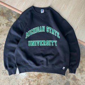 Puede incluir: Sudadera negra de cuello redondo con las palabras "MICHIGAN STATE UNIVERSITY" en letras verdes. La sudadera tiene un ajuste clásico con puños y dobladillo acanalados. Un logotipo de Russell Athletic es visible en la parte superior izquierda del pecho.