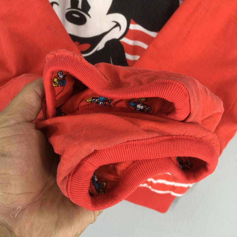 May include: Close-up of a red long-sleeved shirt featuring a large Mickey Mouse graphic. The cuffs are red with a white and red striped design. The inside of the cuffs have a pattern of cartoon characters.