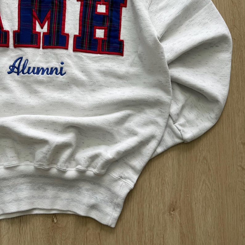 May include: A white heathered sweatshirt featuring large block letters with a blue and red plaid pattern. Below the letters, the word Alumni is embroidered in blue script. The garment has long sleeves, a ribbed hem, and a crew neckline.