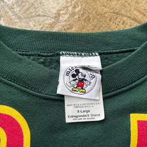 K&ouml;nnte beinhalten: Dunkelgr&uuml;nes X-Large Sweatshirt mit einem Mickey Mouse Logo-Etikett. Das Etikett enth&auml;lt Text auf Englisch, Spanisch und Franz&ouml;sisch. Das Sweatshirt hat rote und gelbe Akzente.
