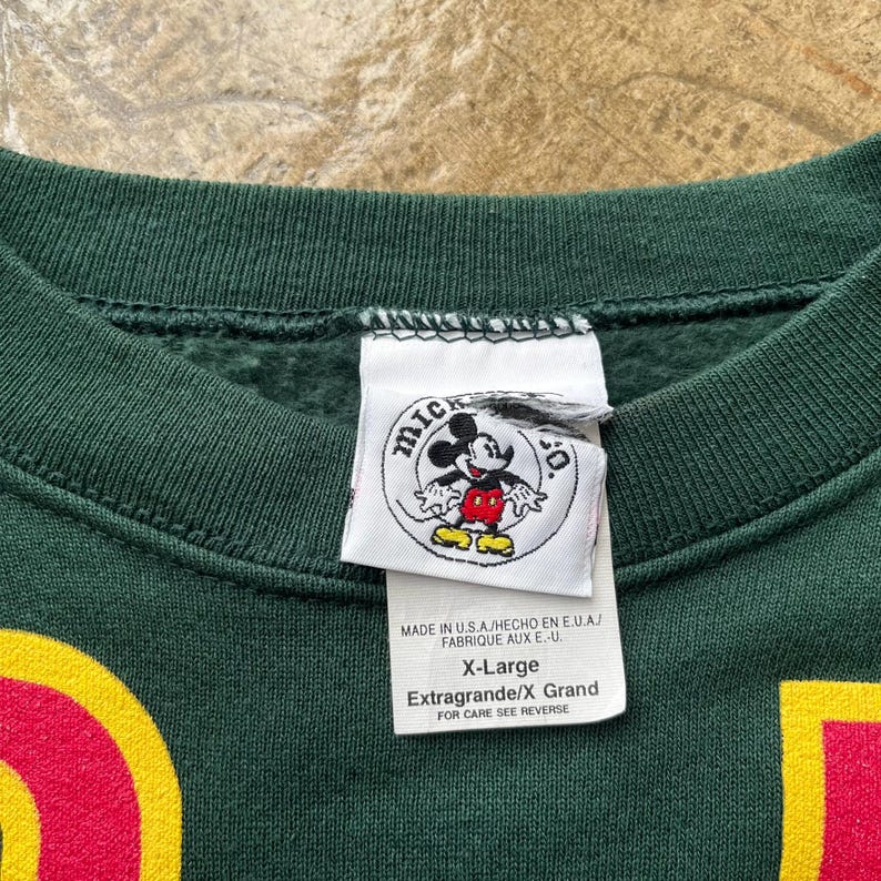 K&ouml;nnte beinhalten: Dunkelgr&uuml;nes X-Large Sweatshirt mit einem Mickey Mouse Logo-Etikett. Das Etikett enth&auml;lt Text auf Englisch, Spanisch und Franz&ouml;sisch. Das Sweatshirt hat rote und gelbe Akzente.