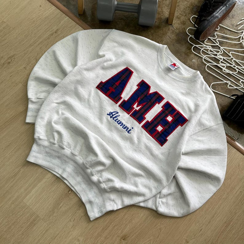 May include: A light gray speckled crewneck sweatshirt featuring the word AME in large, bold, blue and red plaid block letters across the chest. Below the letters, the word Alumni is embroidered in a dark blue script font on the front.
