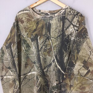 May include: Long-sleeved camouflage shirt in shades of brown, green, and tan, featuring a realistic forest pattern. The shirt is made of a soft material and has a crew neck.