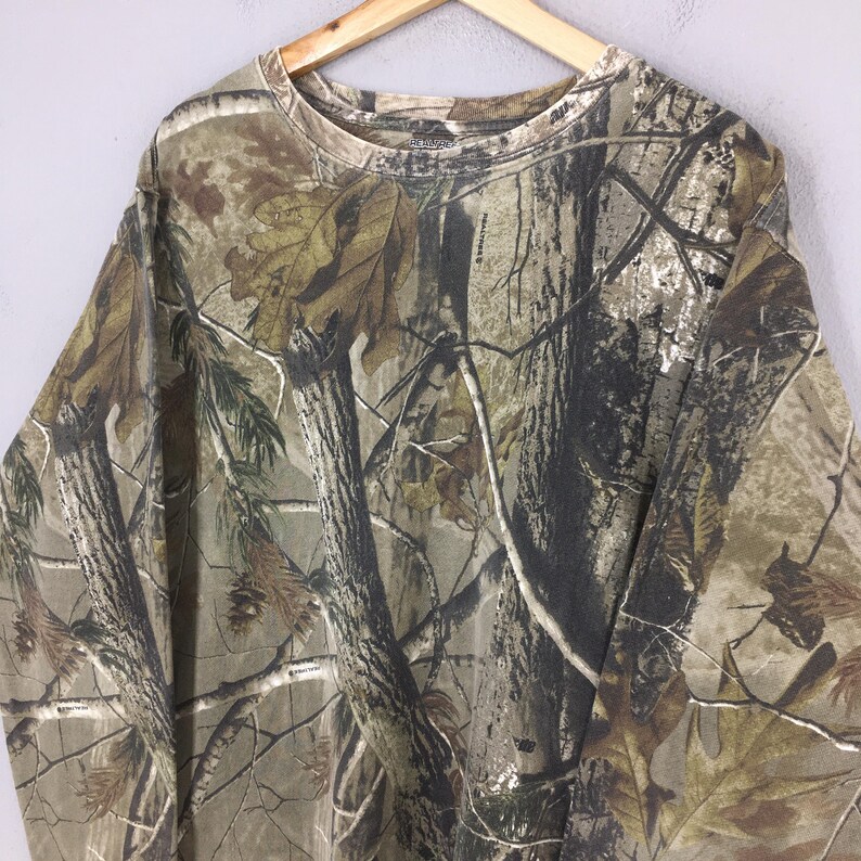 May include: Long-sleeved camouflage shirt in shades of brown, green, and tan, featuring a realistic forest pattern. The shirt is made of a soft material and has a crew neck.