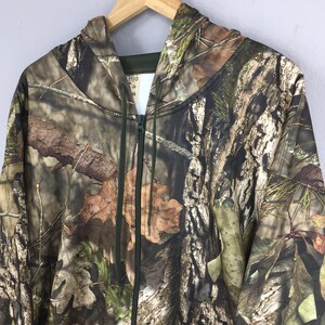 K&ouml;nnte beinhalten: Ein Camouflage-Kapuzenpullover mit Rei&szlig;verschluss, Kapuze und durchgehendem Rei&szlig;verschluss. Das Muster weist eine Mischung aus braunen, gr&uuml;nen und beigen Farben auf, die einer Waldumgebung &auml;hneln. Der Hoodie h&auml;ngt an einem Holzkleiderb&uuml;gel.