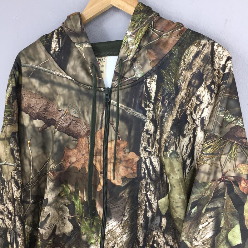 K&ouml;nnte beinhalten: Ein Camouflage-Kapuzenpullover mit Rei&szlig;verschluss, Kapuze und durchgehendem Rei&szlig;verschluss. Das Muster weist eine Mischung aus braunen, gr&uuml;nen und beigen Farben auf, die einer Waldumgebung &auml;hneln. Der Hoodie h&auml;ngt an einem Holzkleiderb&uuml;gel.
