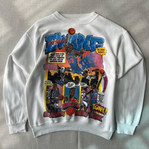 May include: A white long-sleeve sweatshirt featuring a large, colorful graphic print of basketball player Patrick Ewing in action. The design includes comic-style panels, bold blue and red text, and illustrations of basketballs and city skylines.
