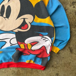 K&ouml;nnte beinhalten: Ein Vintage-Sweatshirt mit einer gro&szlig;en Mickey Mouse-Grafik. Das Sweatshirt hat einen blauen K&ouml;rper mit gelben und roten Akzenten und einen roten unteren Saum. Die &Auml;rmel sind blau mit gelben B&uuml;ndchen.