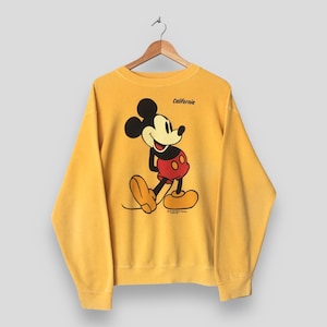 May include: Mustard yellow sweatshirt featuring a large graphic of Mickey Mouse in his classic pose. The cartoon character is wearing red shorts and yellow shoes. The word "California" is printed above the graphic. The sweatshirt is hanging on a wooden hanger.