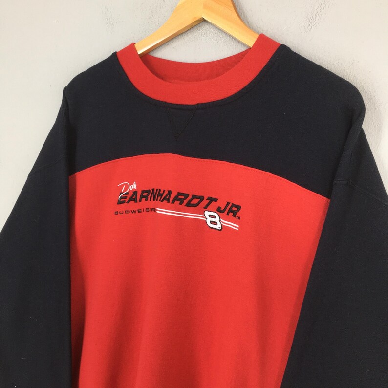 Puede incluir: Una sudadera vintage de cuello redondo en negro, rojo y blanco. La sudadera presenta el texto "Dale Earnhardt Jr." y el n&uacute;mero "8" en blanco. El logotipo de Budweiser tambi&eacute;n es visible. La sudadera est&aacute; colgada en una percha de madera.