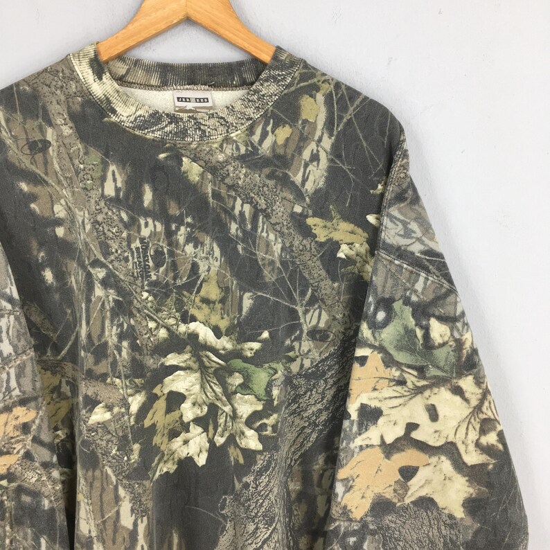 May include: A camouflage-patterned sweatshirt in shades of brown, green, and tan, featuring a crew neck and long sleeves. The fabric displays a detailed print of leaves and branches, creating a woodland effect. The sweatshirt is hanging on a wooden hanger.
