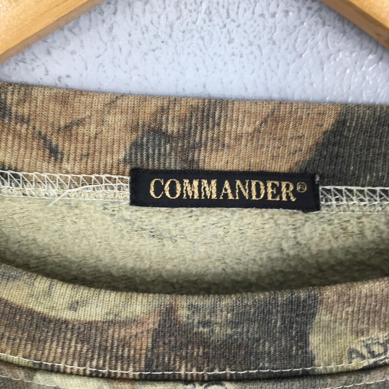 K&ouml;nnte beinhalten: Nahaufnahme eines Sweatshirts mit Tarnmuster. Der Stoff weist Farbt&ouml;ne von Braun, Beige und Gr&uuml;n auf. Ein schwarzes Etikett mit dem Wort "COMMANDER" in goldfarbenen Buchstaben ist auf den Halsausschnitt gen&auml;ht.