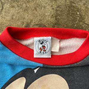 K&ouml;nnte beinhalten: Nahaufnahme eines Vintage-Sweatshirts mit Mickey Mouse. Das Sweatshirt hat einen roten Kragen und ein blau-graues Farbblock-Design. Ein Etikett mit dem Mickey & Co. Logo ist sichtbar.