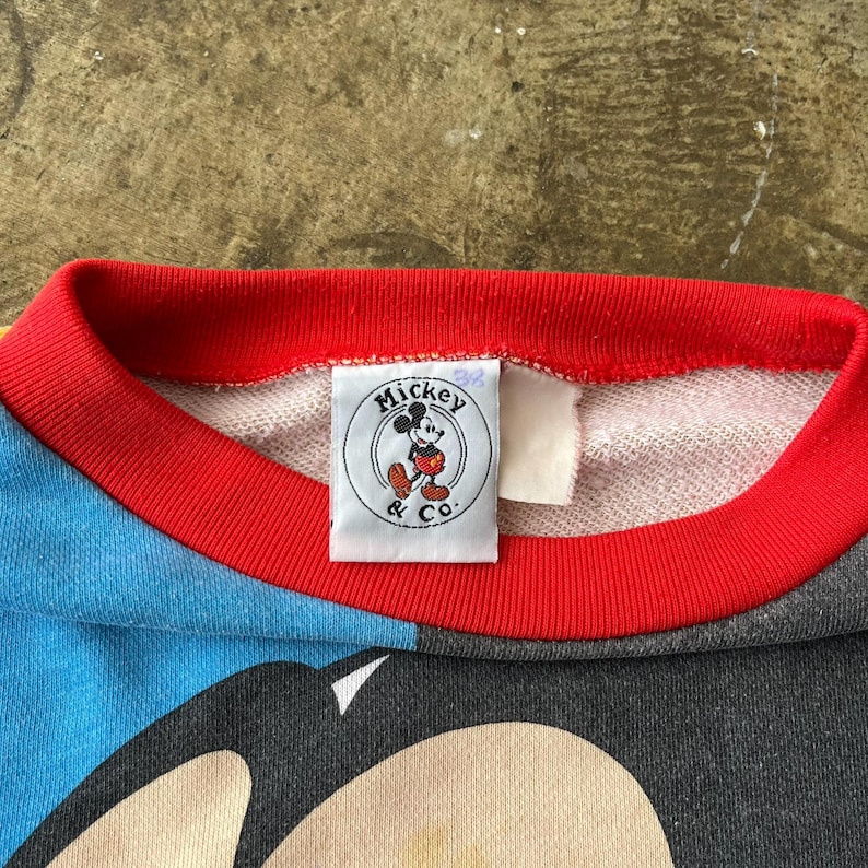 K&ouml;nnte beinhalten: Nahaufnahme eines Vintage-Sweatshirts mit Mickey Mouse. Das Sweatshirt hat einen roten Kragen und ein blau-graues Farbblock-Design. Ein Etikett mit dem Mickey & Co. Logo ist sichtbar.