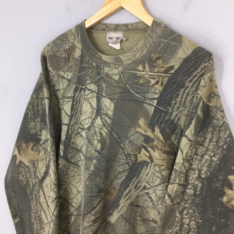 May include: A camouflage-patterned sweatshirt in shades of green, brown, and tan, hanging on a wooden hanger. The long-sleeved top features a crew neck and a dense forest design, suitable for outdoor pursuits.