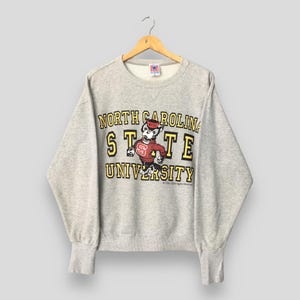 Puede incluir: Una sudadera gris jaspeado de cuello redondo con el texto North Carolina State University en amarillo con bordes negros. Un gr&aacute;fico de una mascota lobo con un su&eacute;ter rojo con NCSU impreso se encuentra centrado en el pecho.