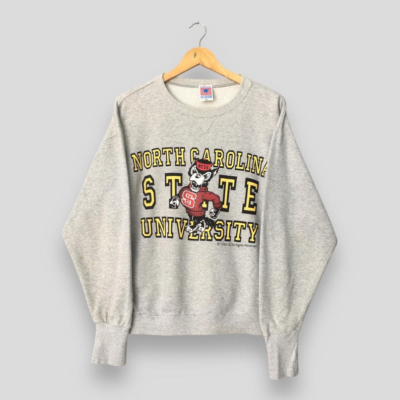 Puede incluir: Una sudadera gris jaspeado de cuello redondo con el texto North Carolina State University en amarillo con bordes negros. Un gr&aacute;fico de una mascota lobo con un su&eacute;ter rojo con NCSU impreso se encuentra centrado en el pecho.
