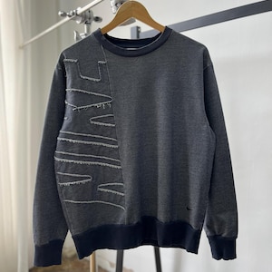 May include: Dark gray sweatshirt with a unique design. The left side features a stitched pattern with exposed seams. The crew neck, cuffs, and hem are a darker shade of gray. A small black logo is visible on the lower right side.