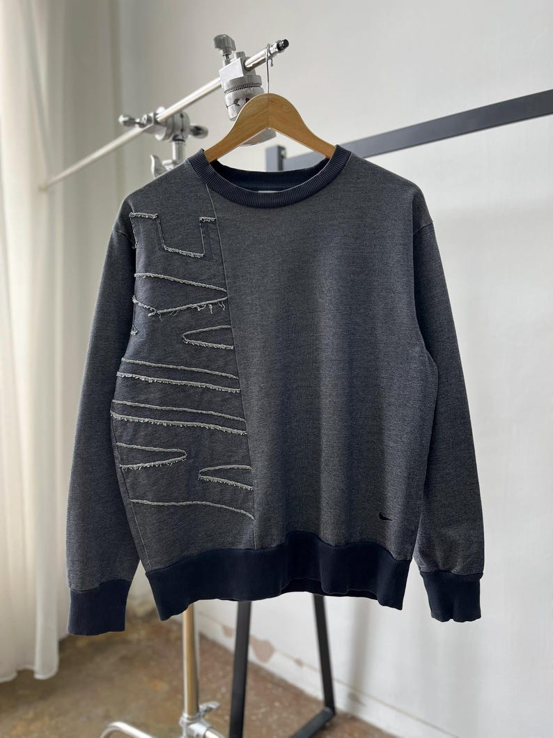 May include: Dark gray sweatshirt with a unique design. The left side features a stitched pattern with exposed seams. The crew neck, cuffs, and hem are a darker shade of gray. A small black logo is visible on the lower right side.