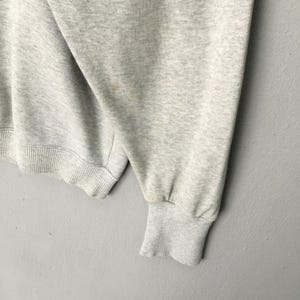 Puede incluir: Una sudadera gris claro jaspeado que presenta una manga larga con un pu&ntilde;o acanalado y un dobladillo acanalado a juego. La tela muestra una textura sutilmente moteada en toda la prenda, con costuras visibles a lo largo de la manga y el dobladillo.