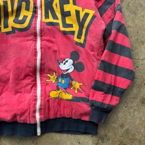May include: A red zip-up jacket featuring Mickey Mouse. The jacket has black and gray striped sleeves and the word "MICKEY" in yellow letters across the top. Mickey Mouse is in the center, with yellow gloves and shoes, blue shorts, and a white face.