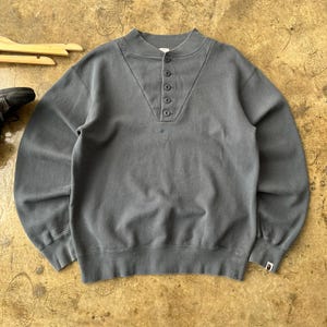 May include: A gray sweatshirt with a button-up collar and long sleeves. The shirt has a small logo on the chest and a ribbed hem and cuffs. The shirt is made of a soft, comfortable material.