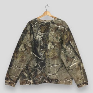 May include: A camouflage-patterned sweatshirt hangs on a wooden hanger. The long-sleeved top features a brown, green, and tan woodland print. The sweatshirt is made of a soft material and has a crew neck.