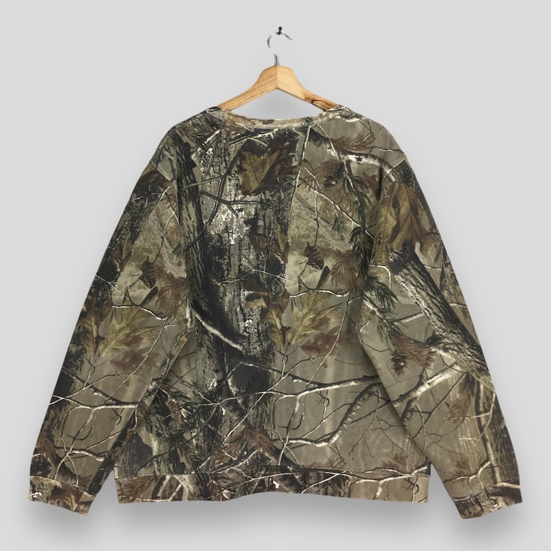 May include: A camouflage-patterned sweatshirt hangs on a wooden hanger. The long-sleeved top features a brown, green, and tan woodland print. The sweatshirt is made of a soft material and has a crew neck.