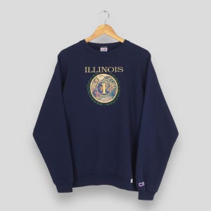 Può includere: Felpa blu navy con la scritta "ILLINOIS" nella parte superiore. Sotto, un emblema circolare con la lettera "I" e le parole "FIGHTING ILLINI". La felpa ha maniche lunghe e girocollo.